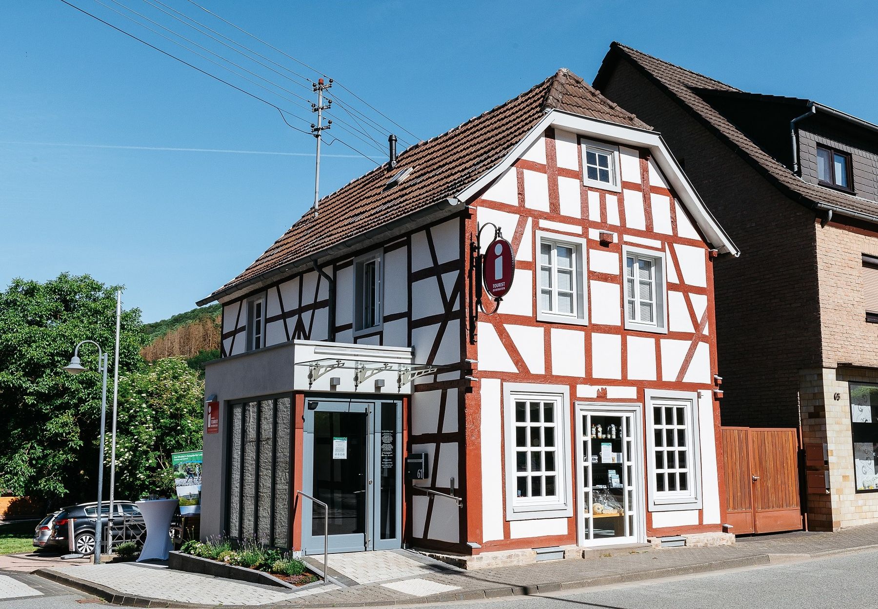 A traditional half-timbered building with white walls and brown wooden beams stands on a sunny street next to modern and brick buildings. There is a small glass entrance on the left side and a red sign above the door.